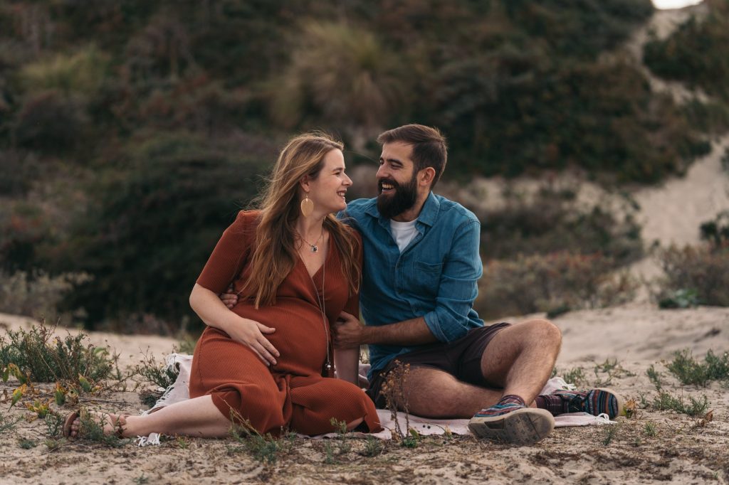 Pregnant woman and her partner sitting together in the dunes during an outdoor maternity photoshoot in the Netherlands