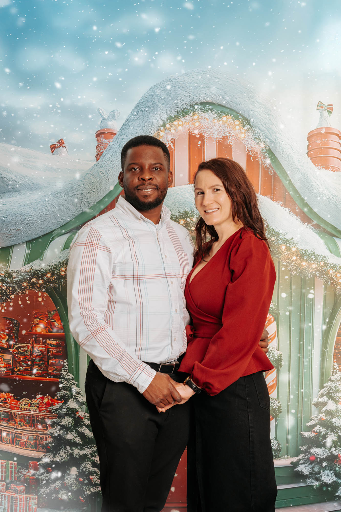 Couple posing in front of festive Christmas gingerbread house backdrop with snow effect during holiday photography session