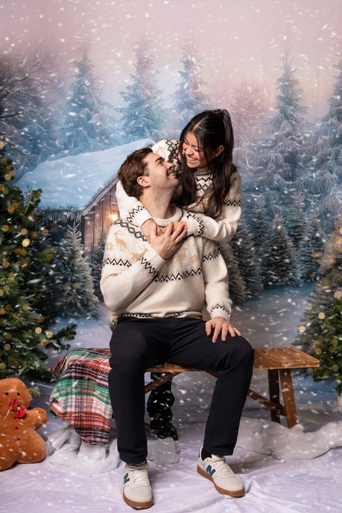 Couple in matching cream Christmas sweaters embracing and laughing on a bench in a snowy forest backdrop