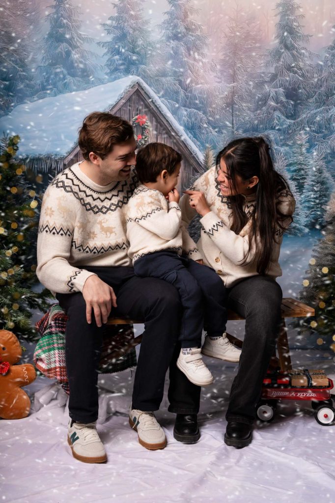 Family of three with a baby in festive sweaters, sitting beside a large teddy bear in a snowy forest backdrop