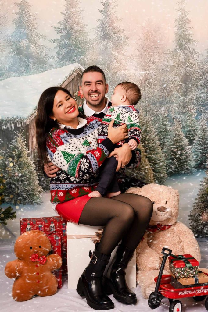 Family of three in matching cream Christmas sweaters laughing together on a bench in front of a snowy cabin backdrop