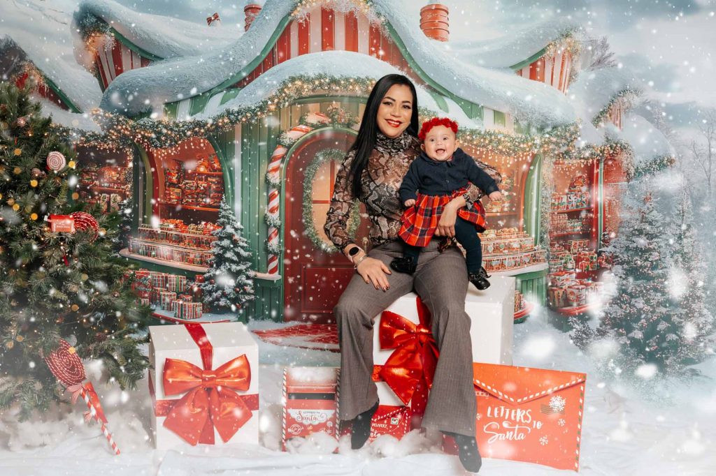 Mother smiling with her baby girl in a red plaid dress, seated in front of Santa's workshop Christmas backdrop