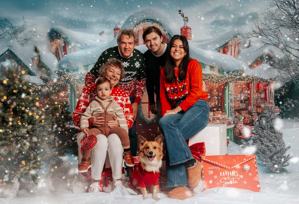 Multigenerational family with grandparents, parents, toddler, and dog posing together in front of Santa's workshop Christmas backdrop