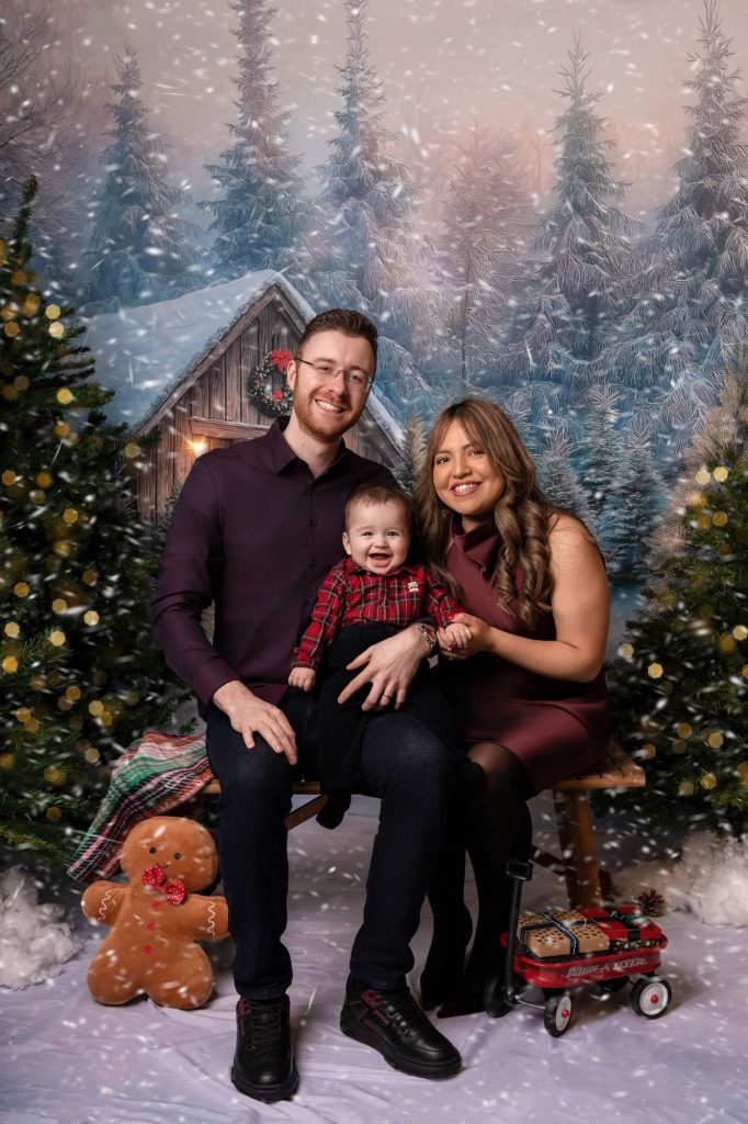 Parents smiling with their baby boy on a bench in front of a snowy wooden cabin Christmas backdrop