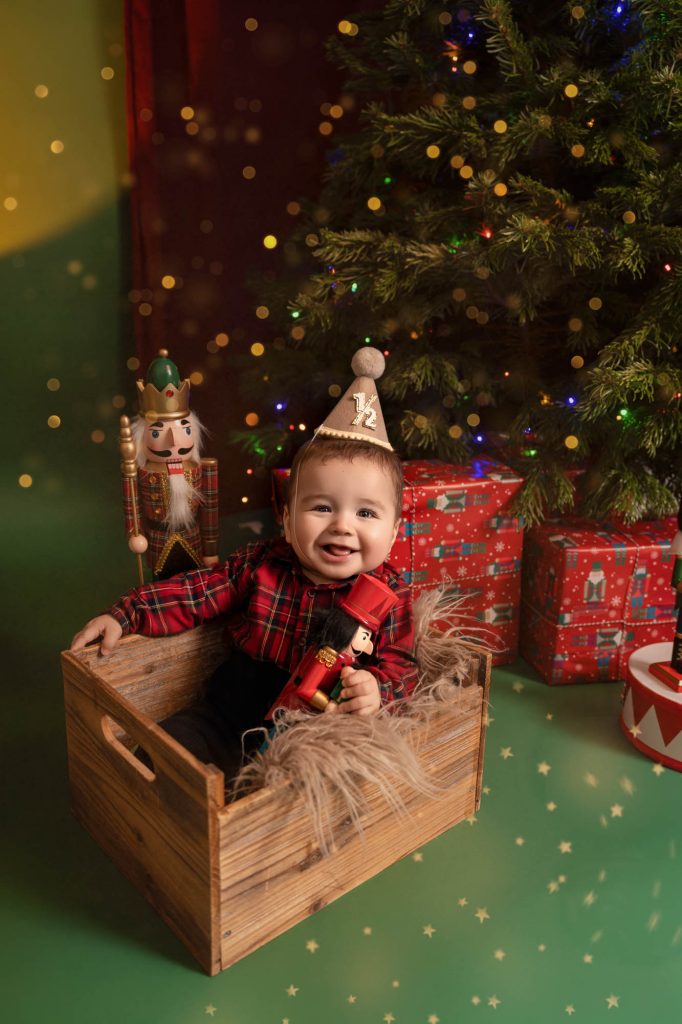 Baby boy in plaid shirt wearing a half-birthday hat sitting in a wooden crate next to a nutcracker toy, Christmas tree in background