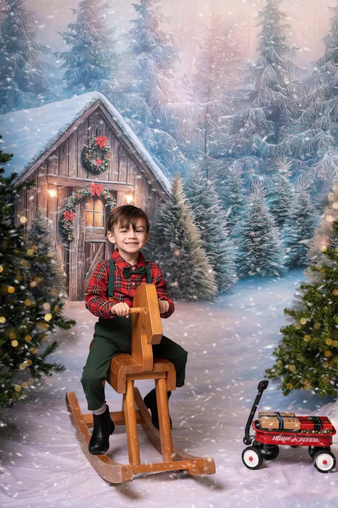 Boy in plaid shirt and bow tie riding a wooden rocking horse in front of a snowy wooden cabin Christmas backdrop