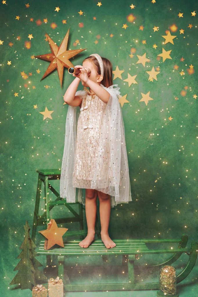 Girl in shimmering gold dress and cape looking through a toy telescope against a green starry Christmas backdrop