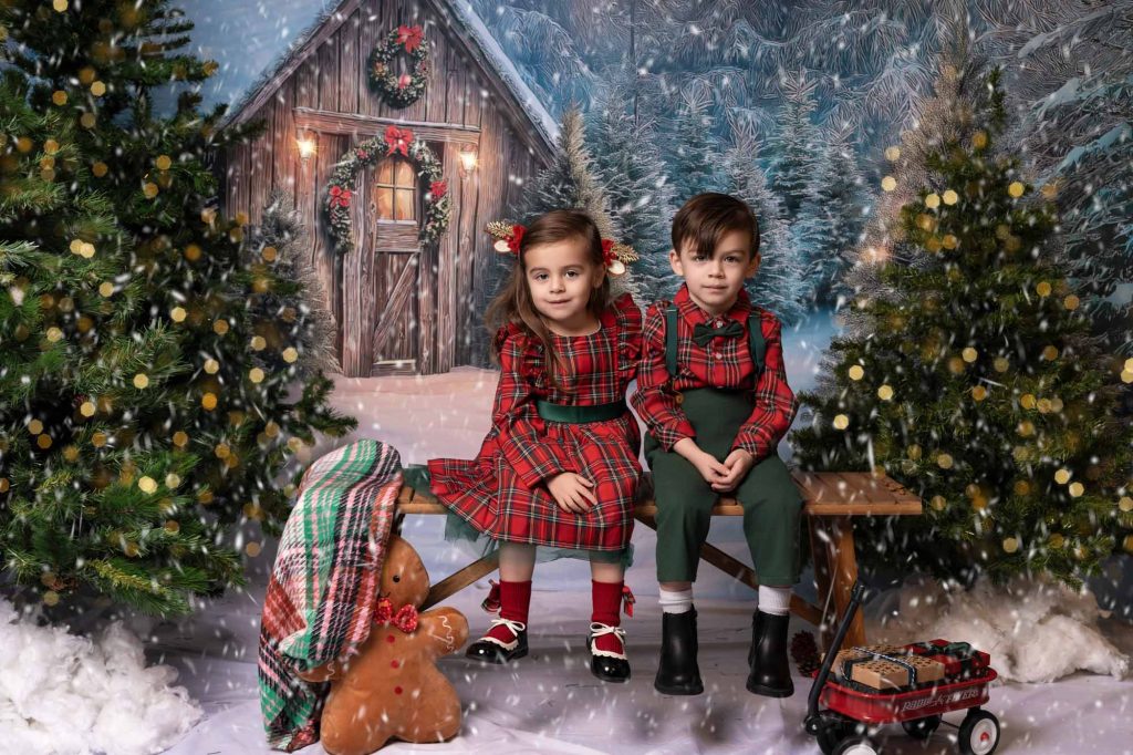 Young girl and boy in coordinating beige holiday outfits sitting together, festive green and red backdrop with Christmas tree
