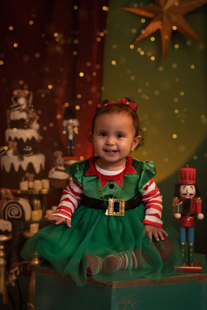 Toddler girl in a green and red elf-style dress sitting on a green box, gold star and nutcracker in background