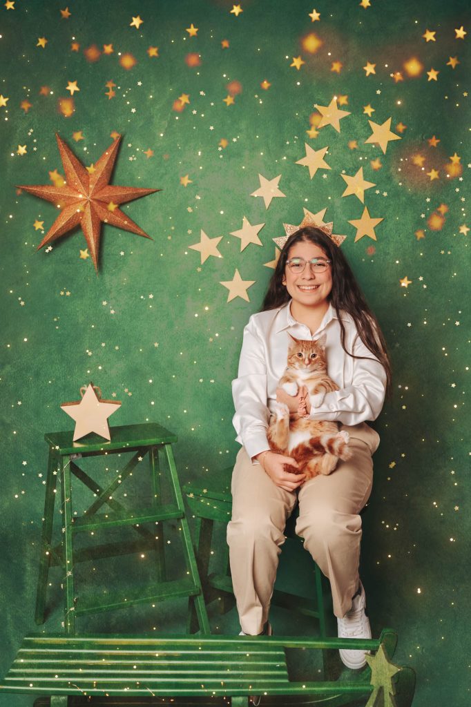 Teen girl wearing a star headband smiling while holding an orange tabby cat, green starry Christmas backdrop