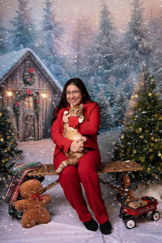 Woman in a red suit smiling while holding an orange tabby cat on a bench in a snowy Christmas forest backdrop