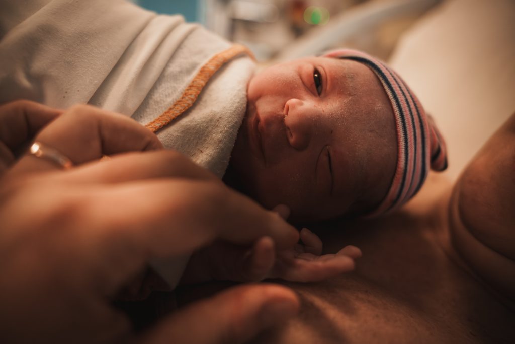 Newborn baby with striped hat resting skin-to-skin on mother's chest moments after birth, father's hand holding baby's tiny hand