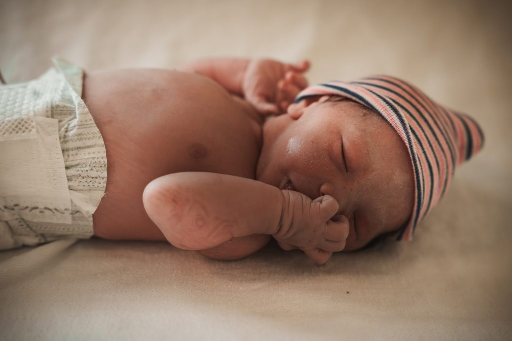Newborn baby sleeping peacefully with hand near face, wearing a striped hospital hat